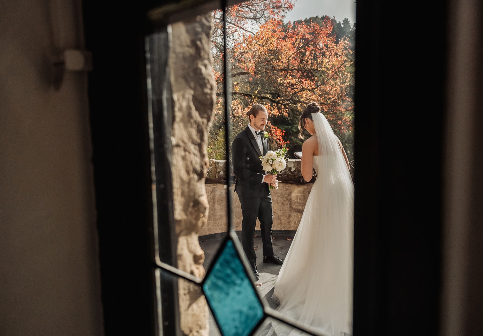 the manor wedding - crystal & mackenzie 43 Bride and groom on autumn wedding terrace framed by rustic stone window.