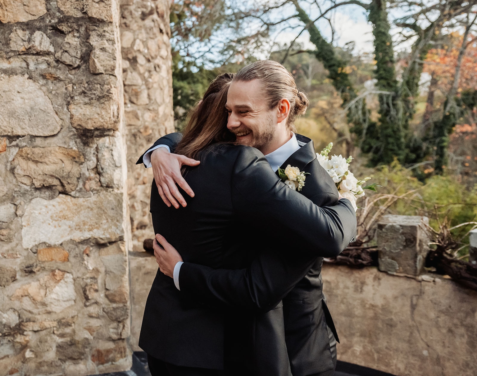the manor wedding - crystal & mackenzie 35 Joyful wedding couple embracing outdoors by a rustic stone wall.