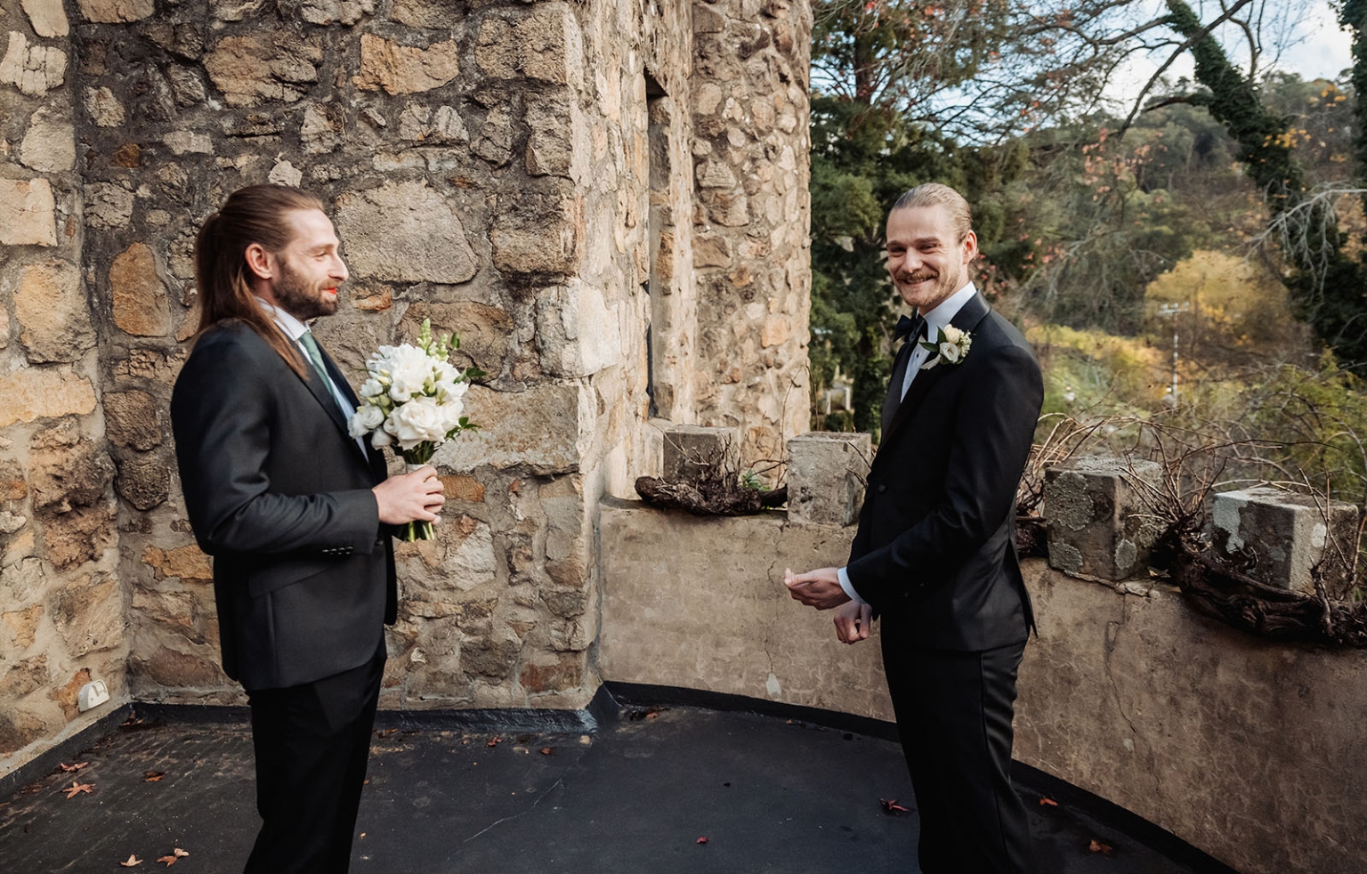 the manor wedding - crystal & mackenzie 34 Two grooms share a joyful wedding moment on a rustic stone terrace.