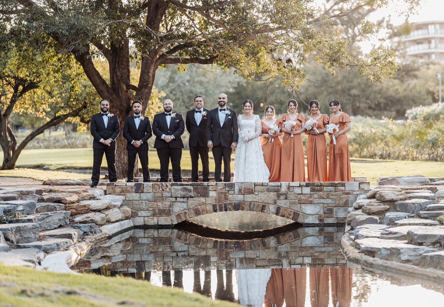st george & st shenouda coptic church wedding - carmina & bill 62 Wedding party portrait on stone bridge in garden with bride, groom, bridesmaids, and groomsmen.