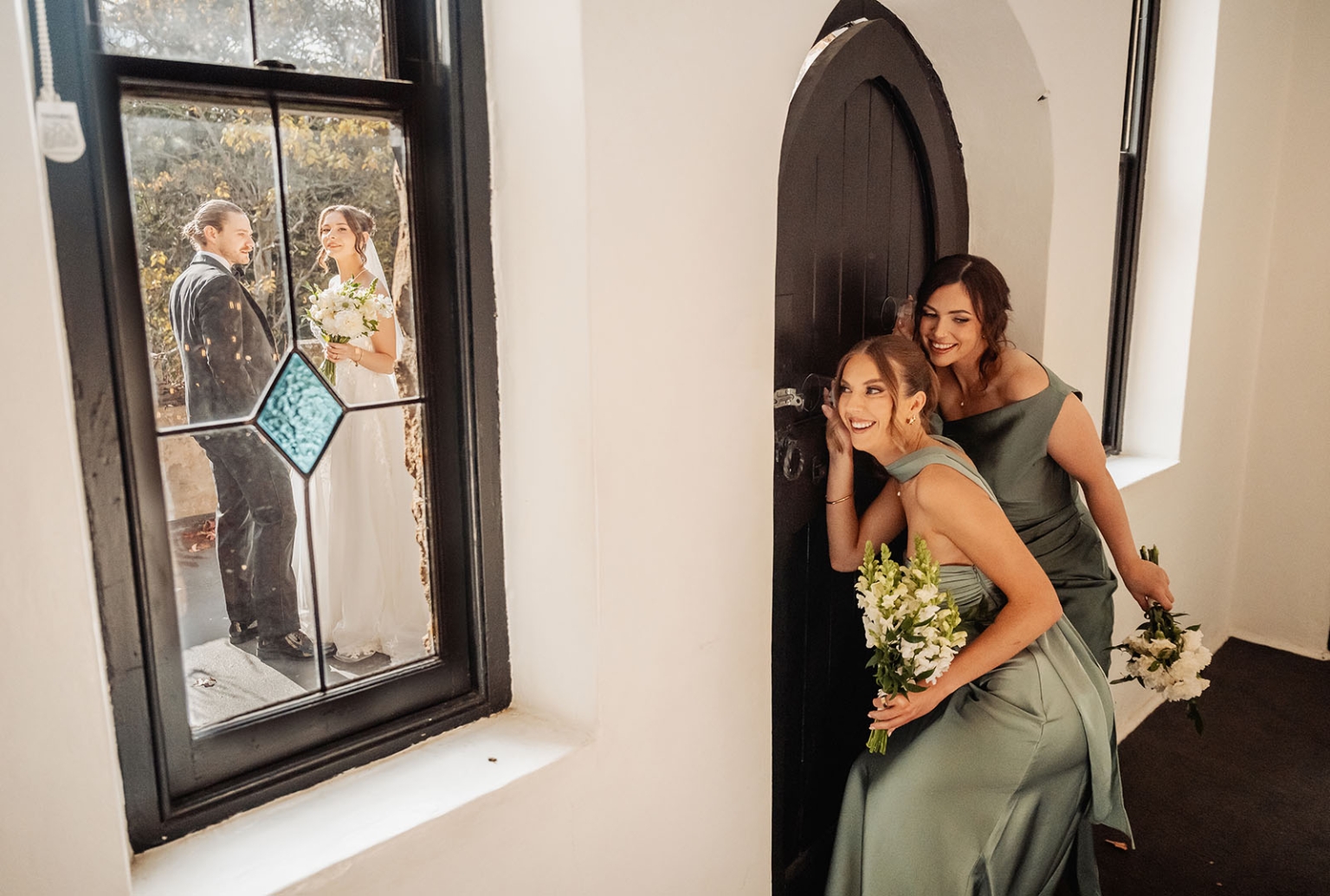 the manor wedding - crystal & mackenzie 44 Bridesmaids peek through chapel door as bride and groom pose outside window.