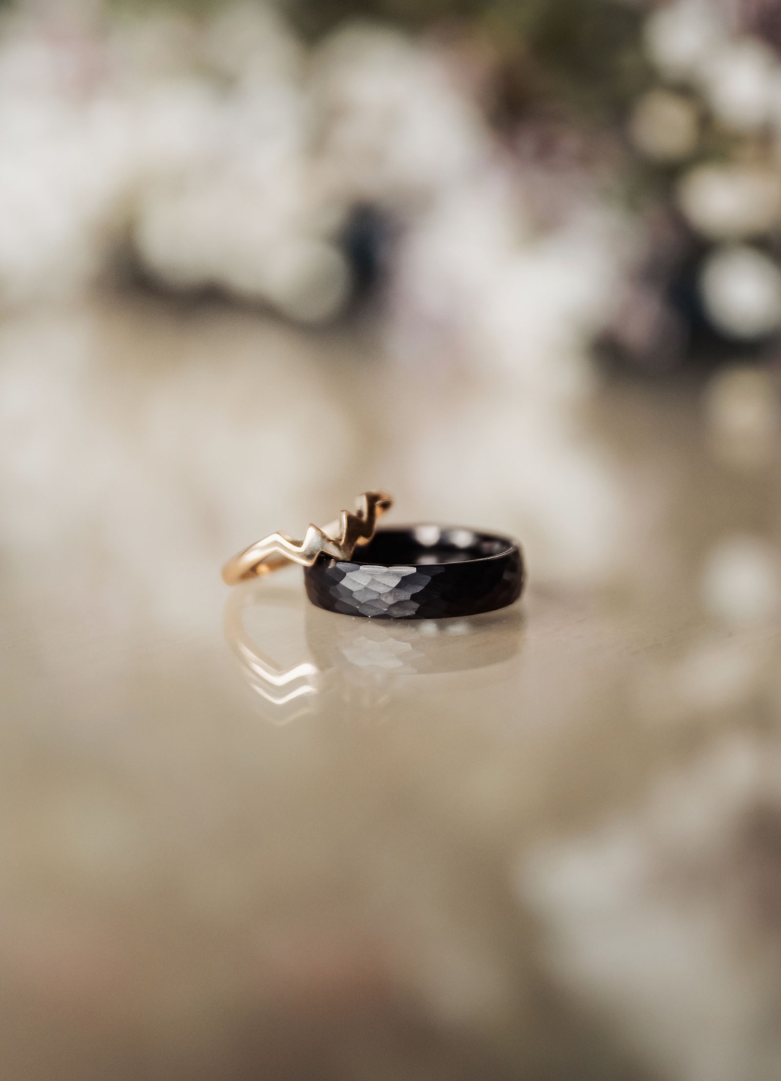 basket range wedding at the manor - leah & wes 1 Black hammered wedding band and gold crown ring on reflective surface.