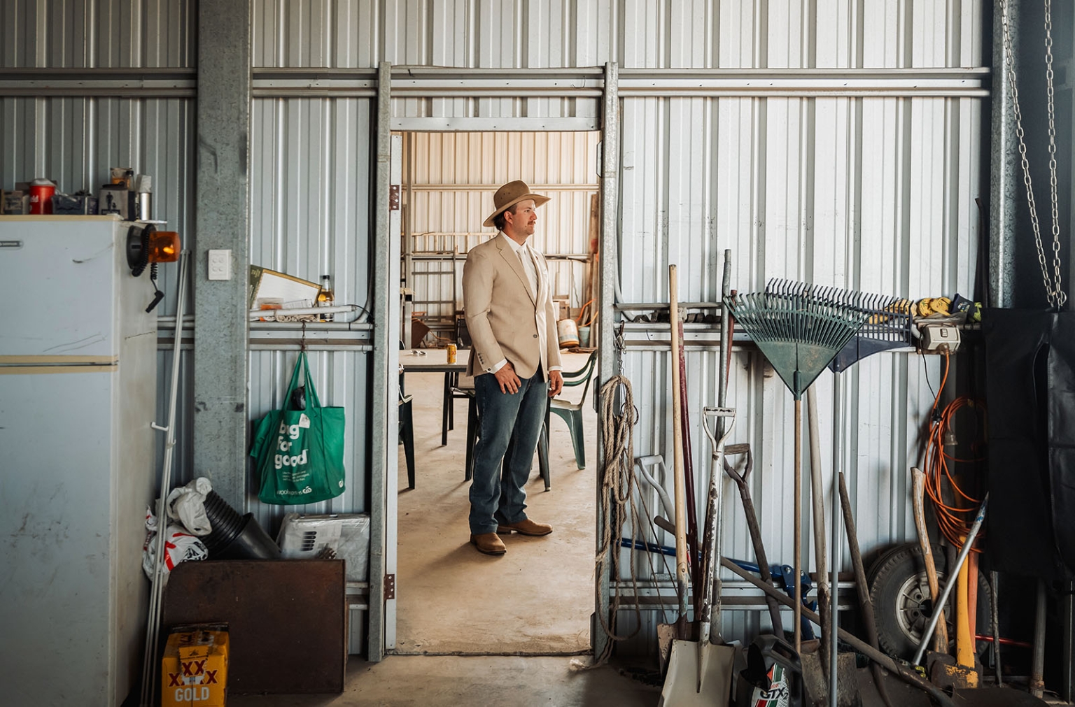 capella wedding photography – caitlin & adam’s farm wedding 118 Adam wearing a cream jacket and cowboy hat stands in the doorway of a farm shed beside gardening tools.