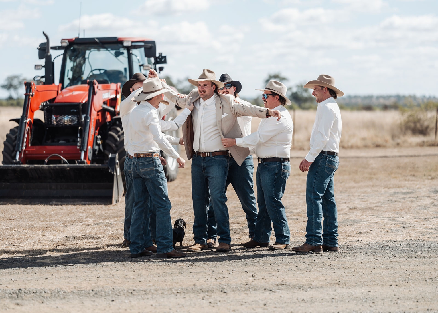 capella wedding photography – caitlin & adam’s farm wedding 114 Adam and his groomsmen in cowboy hats adjust his jacket beside a red tractor on the farm, with a small dog at their feet.
