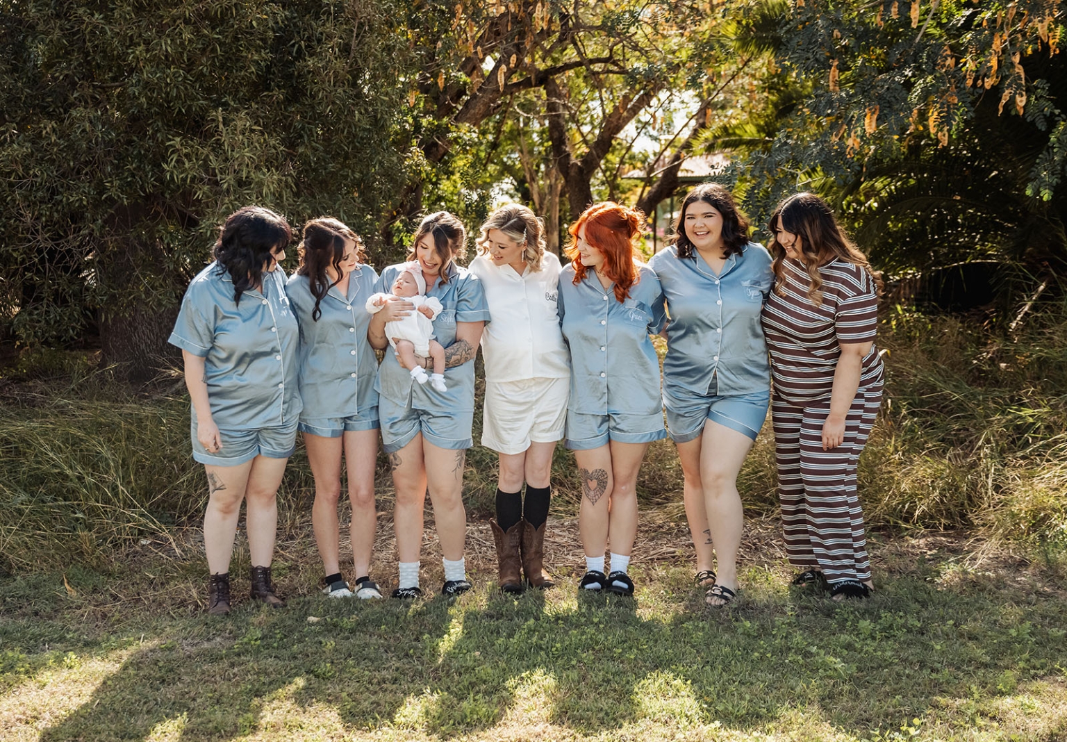 capella wedding photography – caitlin & adam’s farm wedding 124 Seven women, six in matching light blue pyjamas, stand with a baby beneath trees at a farm wedding.