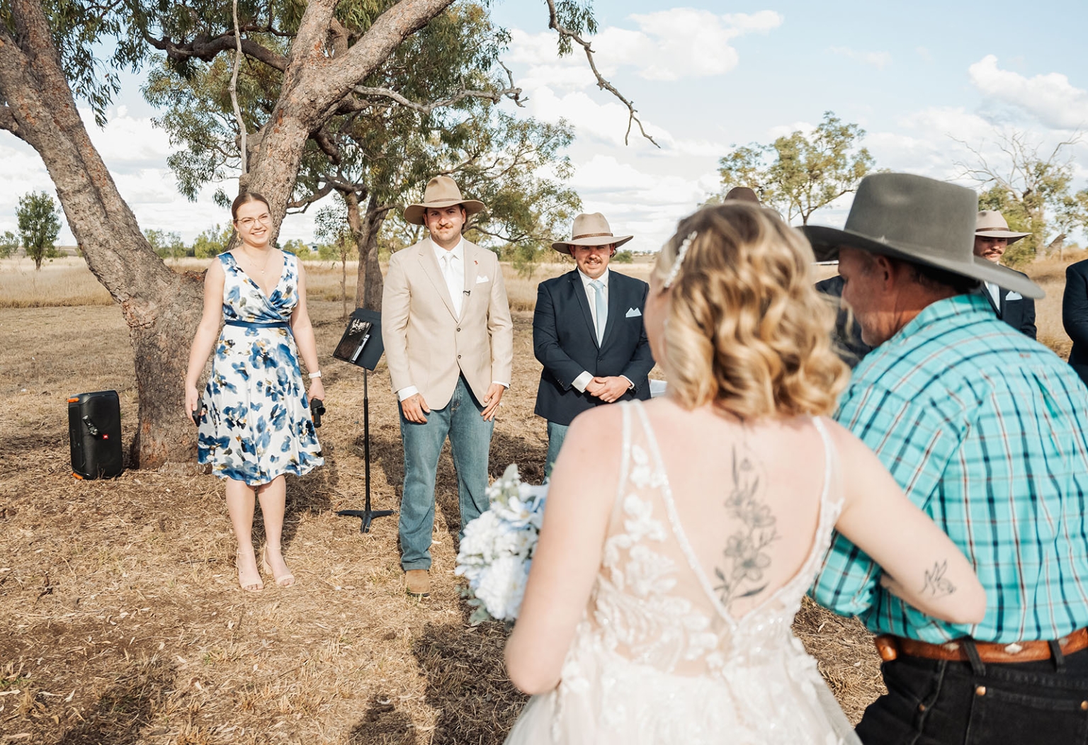 capella wedding photography – caitlin & adam’s farm wedding 139 Bride in a lace wedding dress walking towards the ceremony with an older man in a cowboy hat, as the groom and guests wait beneath a tree.