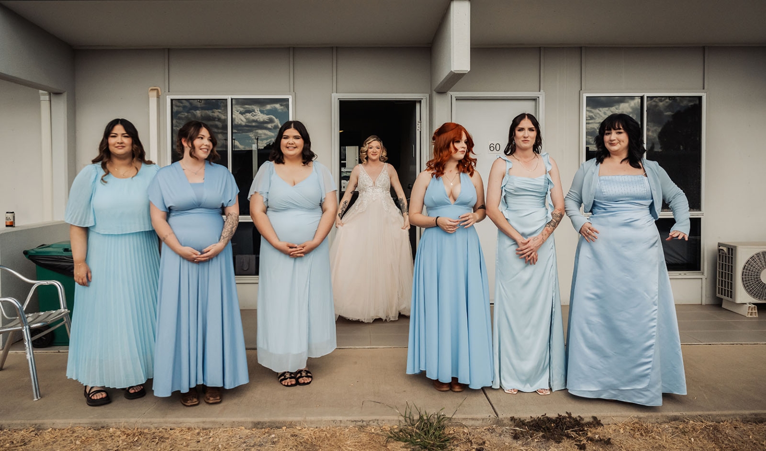 capella wedding photography – caitlin & adam’s farm wedding 129 Bride in a white wedding gown standing behind six bridesmaids in light blue dresses outside a house