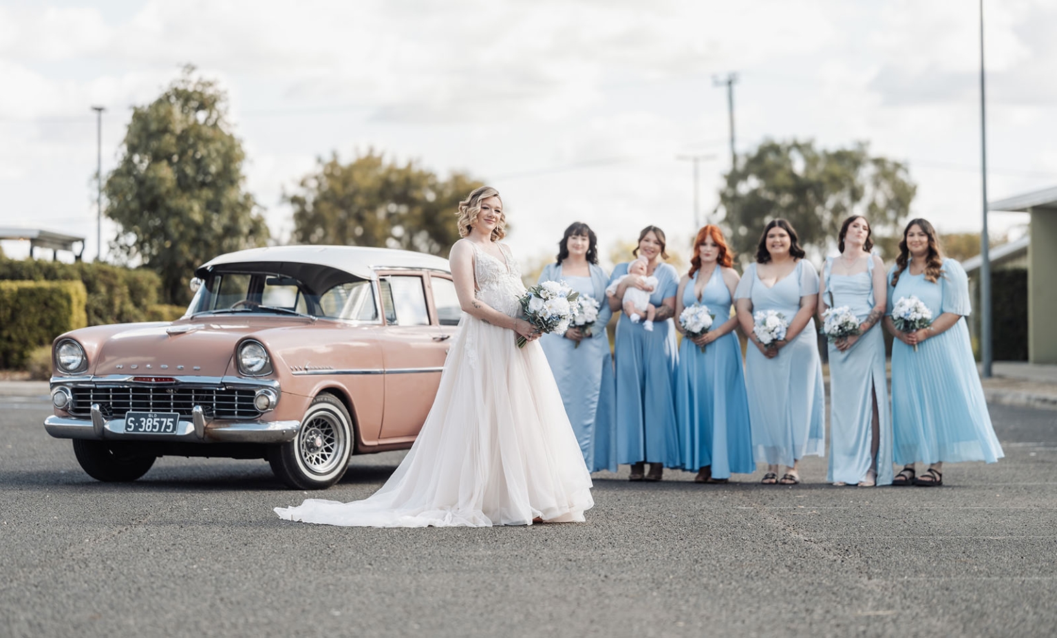 capella wedding photography – caitlin & adam’s farm wedding 133 Bride holding a white bouquet beside a vintage pink Dodge car, with bridesmaids in pale blue dresses behind her