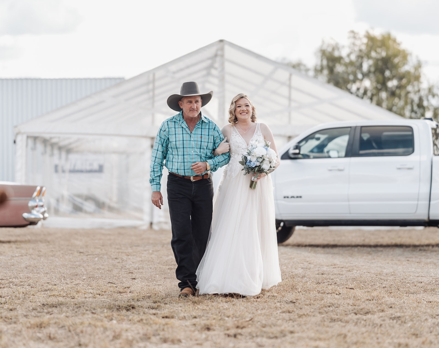 capella wedding photography – caitlin & adam’s farm wedding 138 Bride in a white gown carrying a bouquet walks arm in arm with a man in a cowboy hat outside a marquee.