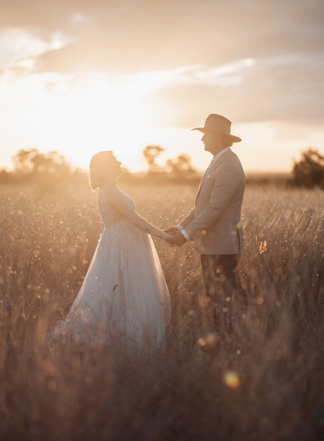 capella wedding photography – caitlin & adam’s farm wedding 170 Caitlin and Adam holding hands in a grassy field at sunset, with Adam wearing a wide-brimmed hat and Caitlin in a lace wedding dress.