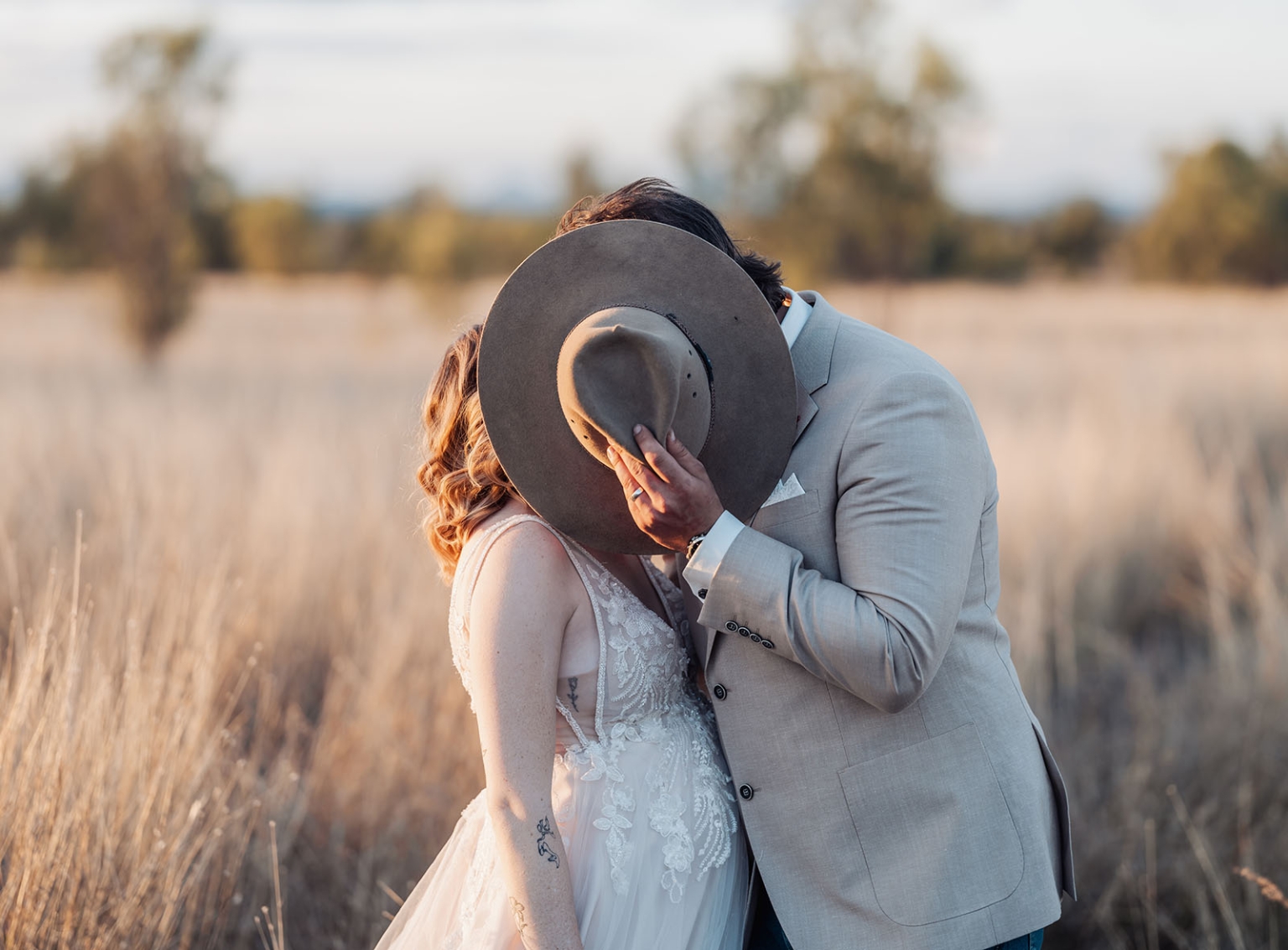capella wedding photography – caitlin & adam’s farm wedding 171 Caitlin and Adam kissing in a sunlit field, with Adam holding his wide-brimmed hat in front of their faces