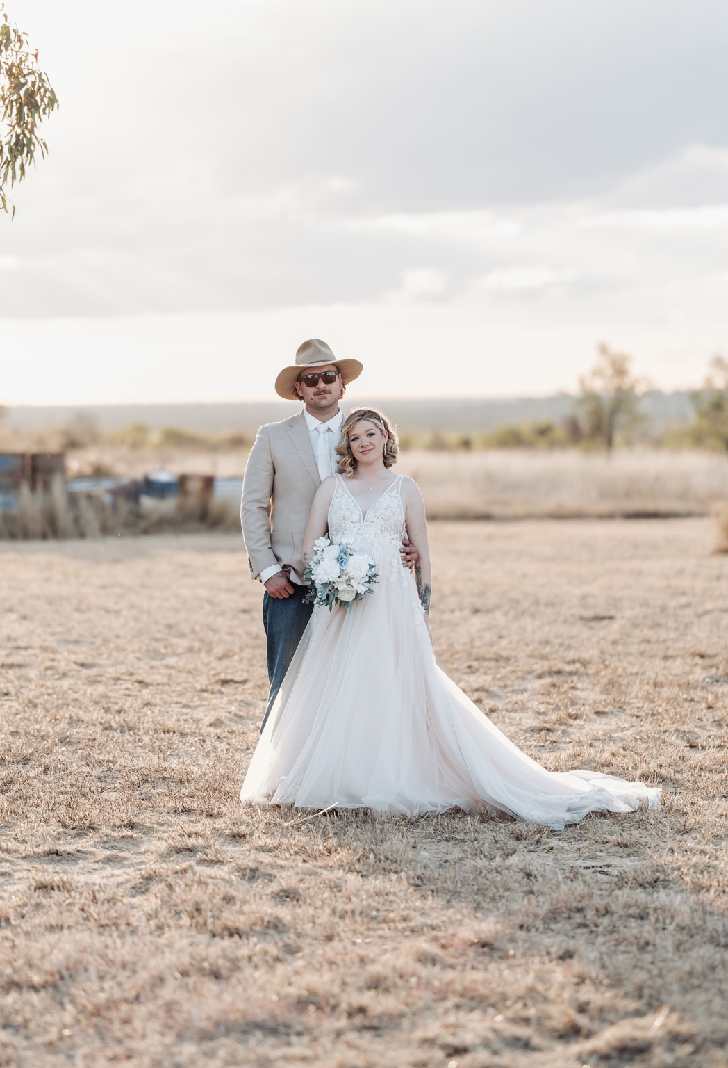 capella wedding photography – caitlin & adam’s farm wedding 165 Bride in a white gown holding a bouquet with groom in a beige jacket and cowboy hat on a dry farm field in Capella