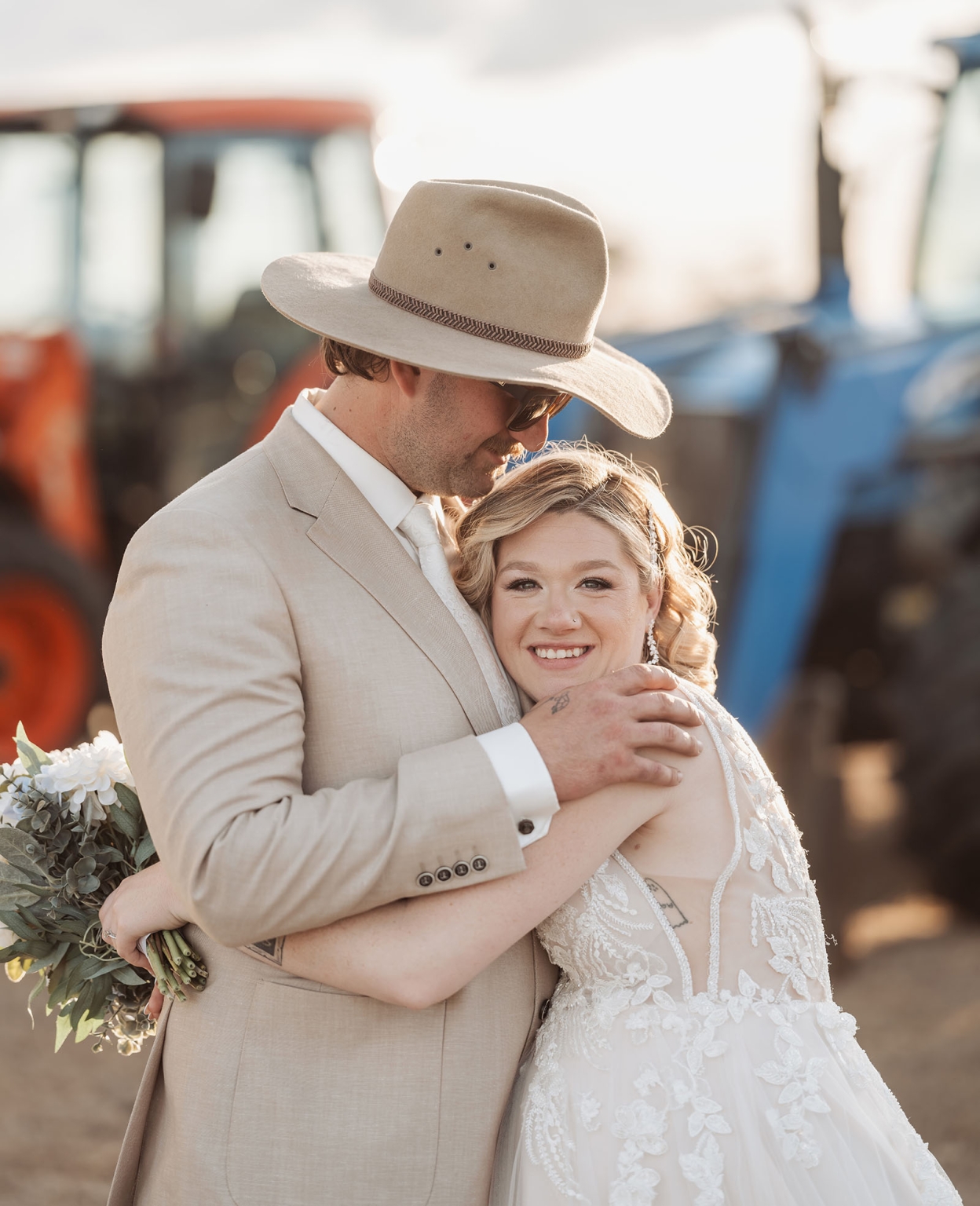 capella wedding photography – caitlin & adam’s farm wedding 162 Caitlin and Adam embracing at their farm wedding, with Adam wearing a wide-brim hat and Caitlin holding a white floral bouquet beside a tractor.