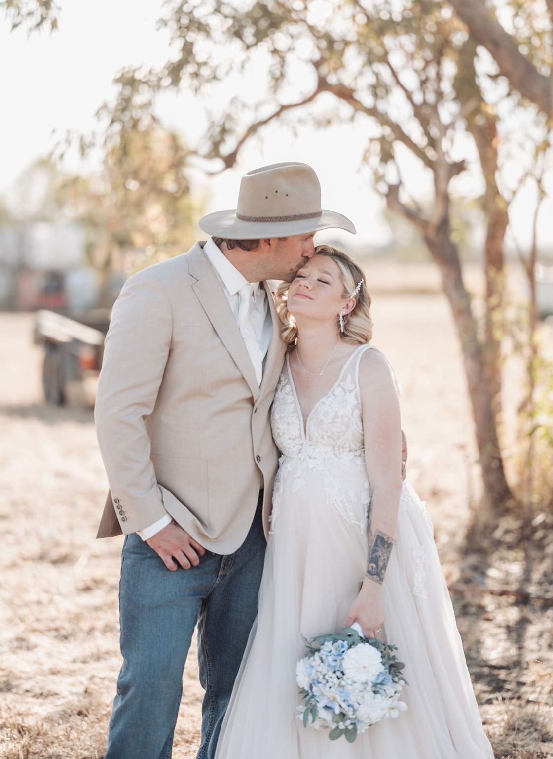 capella wedding photography – caitlin & adam’s farm wedding 151 Adam in a cowboy hat kisses Caitlin on the forehead as she holds a blue and white bouquet beneath a tree at their farm wedding.