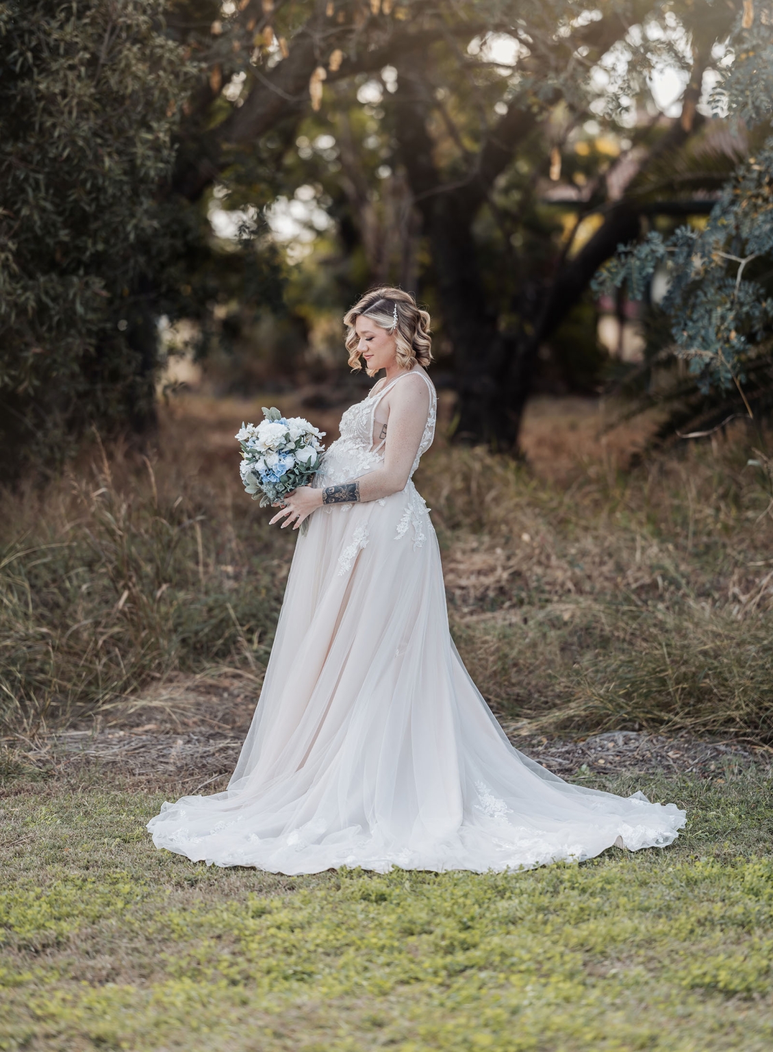 capella wedding photography – caitlin & adam’s farm wedding 135 Caitlin in a lace wedding dress holding a white and blue bouquet in a tree-lined farm setting in Capella.