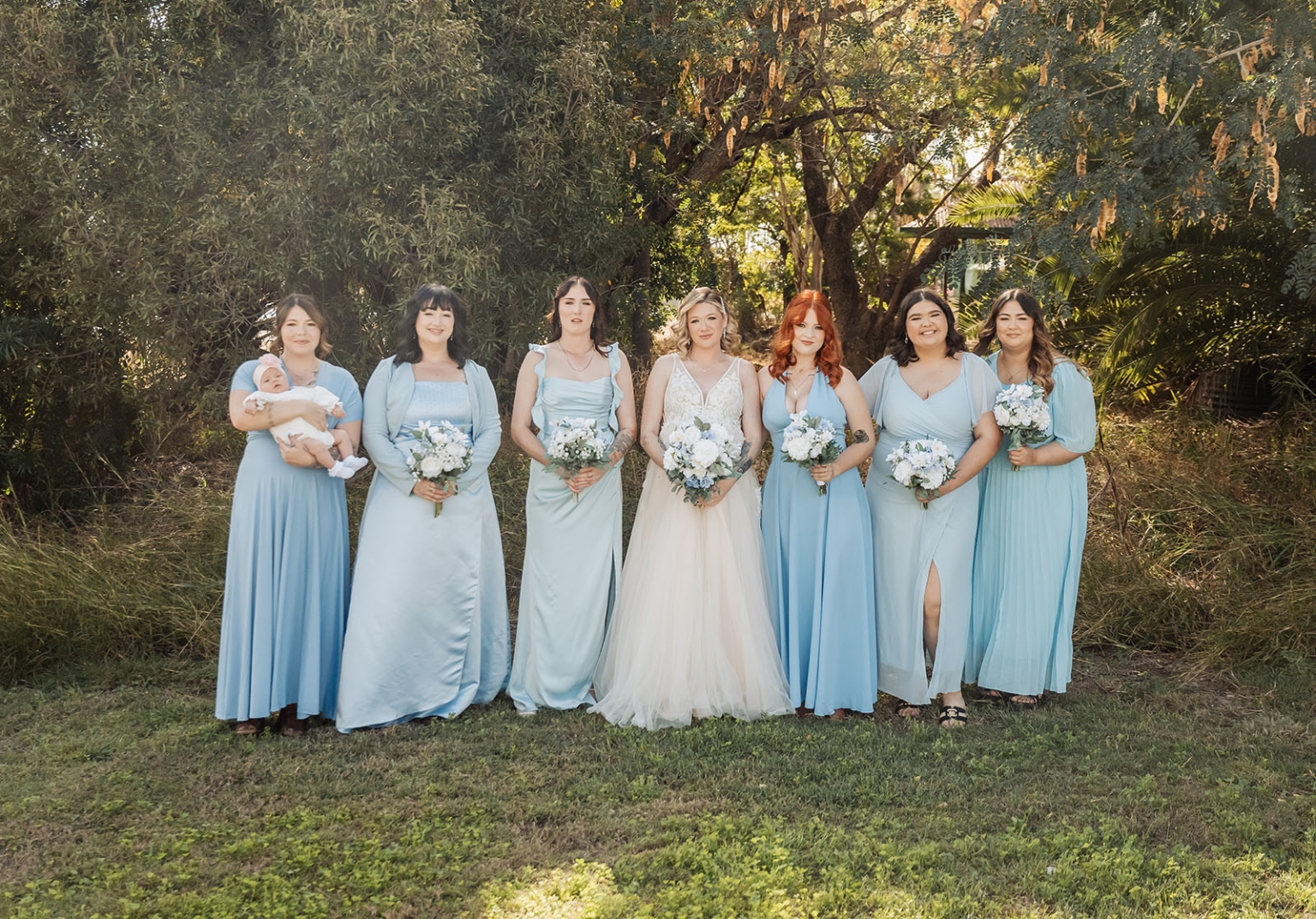 capella wedding photography – caitlin & adam’s farm wedding 134 Bride Caitlin standing with six bridesmaids in pale blue dresses, one holding a baby, beneath leafy trees at a Capella farm wedding