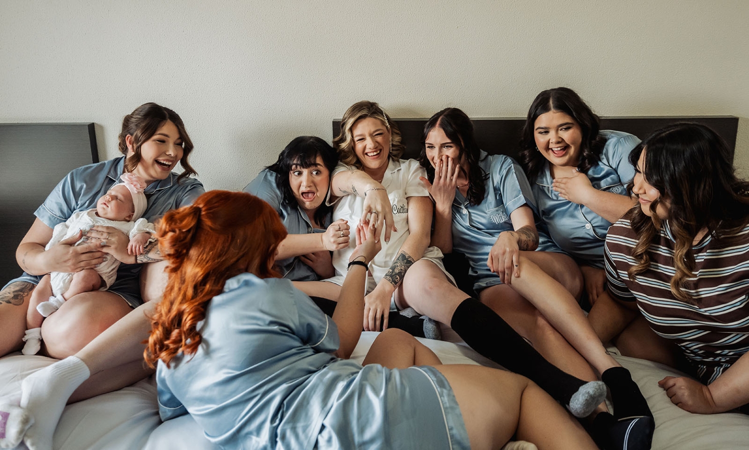 capella wedding photography – caitlin & adam’s farm wedding 125 Caitlin laughing with bridesmaids in matching blue pyjamas on a bed, alongside a woman holding a baby