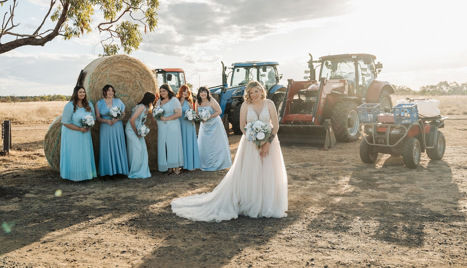 capella wedding photography – caitlin & adam’s farm wedding 152 Bride in a white gown with bridesmaids in pale blue dresses beside hay bales and tractors at a farm wedding