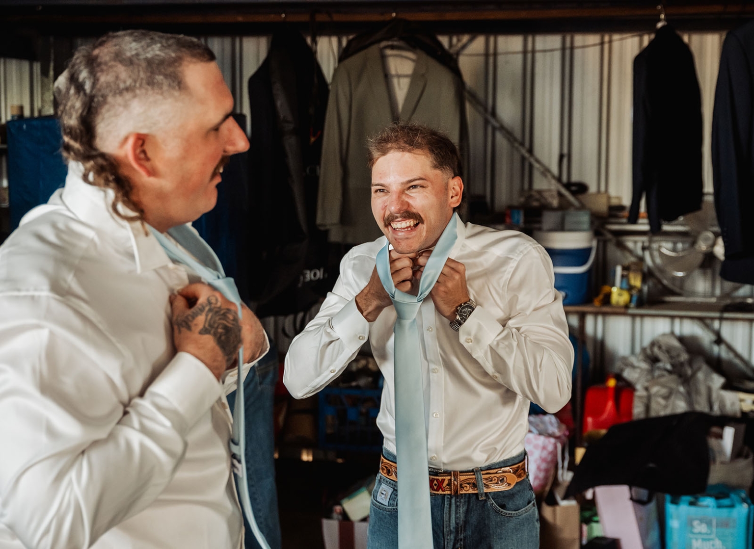 capella wedding photography – caitlin & adam’s farm wedding 116 Two men in white shirts laughing as they adjust pale blue neckties during wedding preparations at a farm in Capella.