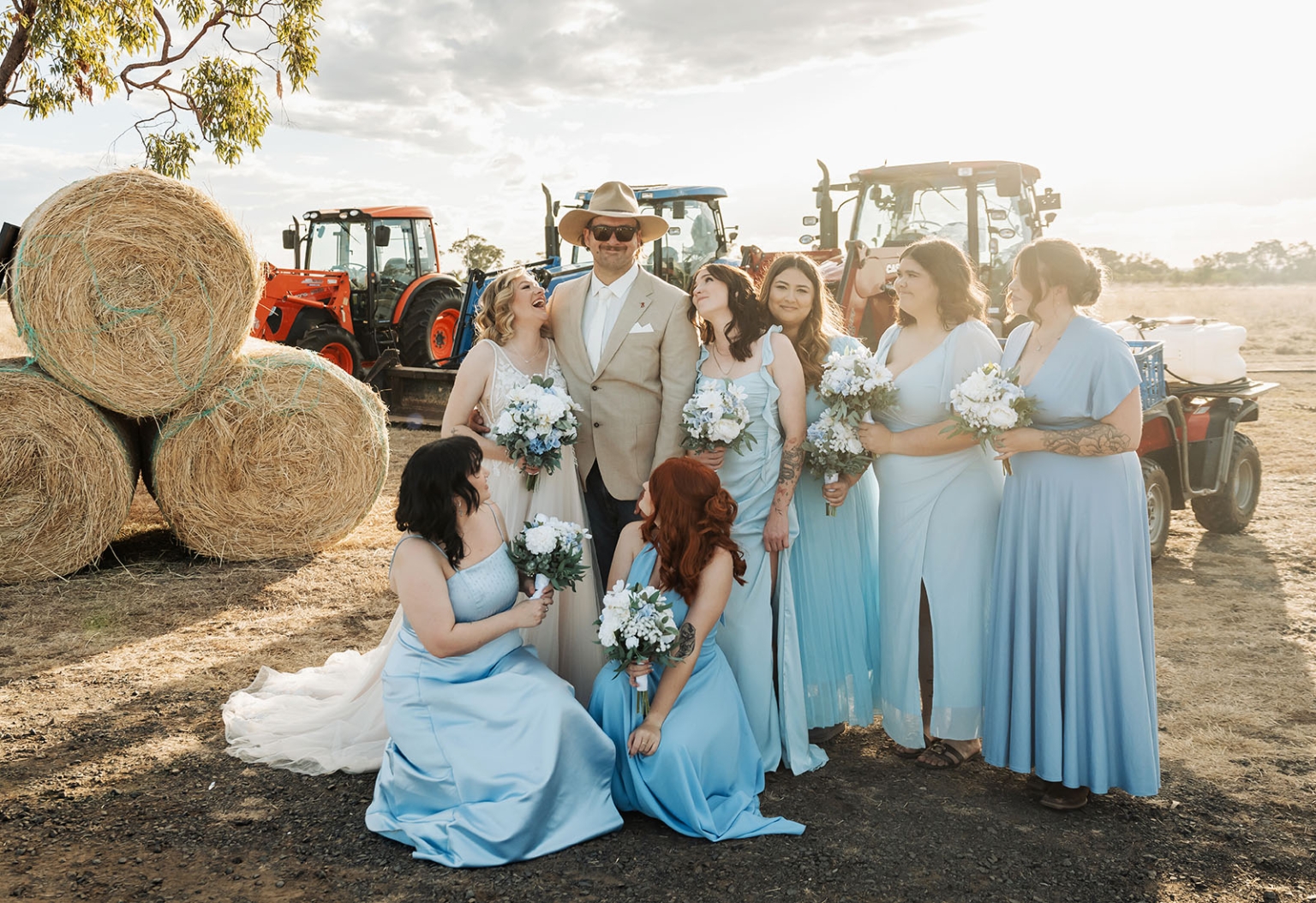 capella wedding photography – caitlin & adam’s farm wedding 153 Bride and groom with six bridesmaids in blue dresses beside hay bales and tractors at a Capella farm wedding