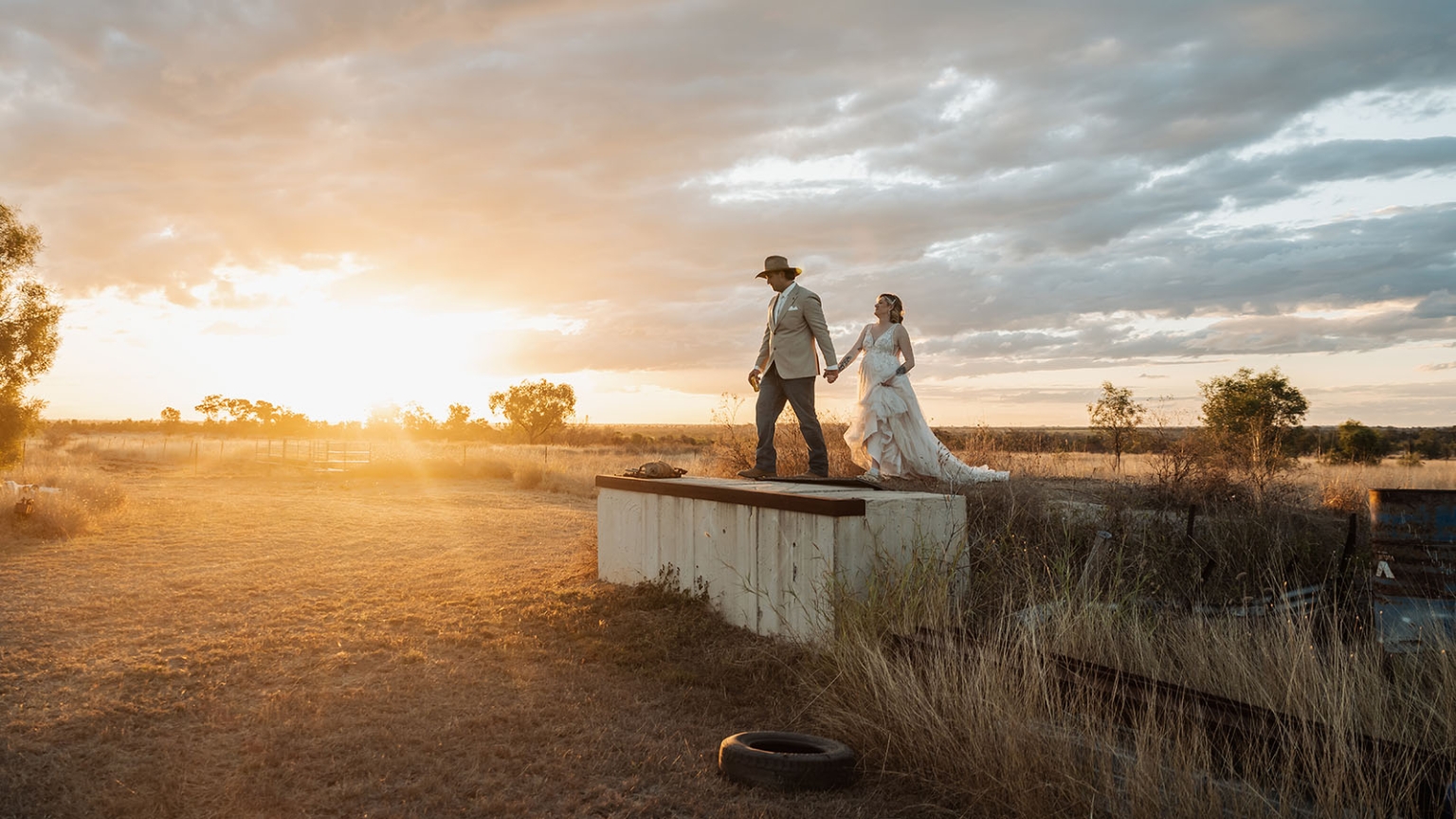 capella wedding photography – caitlin & adam’s farm wedding 173 Bride and groom holding hands as they walk across a farm water tank at sunset in Capella, Queensland