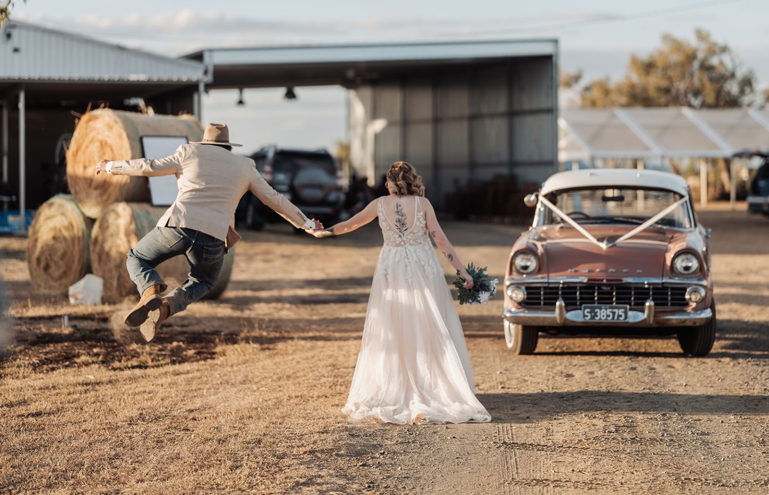 capella wedding photography – caitlin & adam’s farm wedding 160 Newlywed couple holding hands at a farm wedding as the groom jumps beside a decorated vintage car