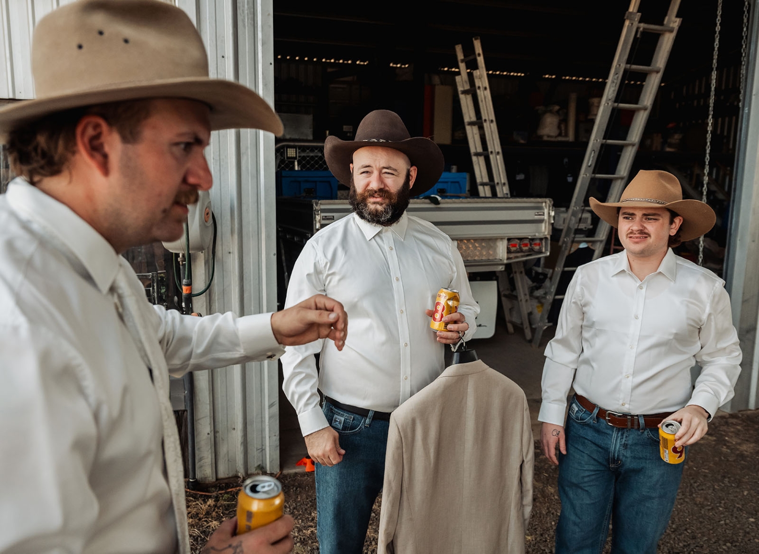 capella wedding photography – caitlin & adam’s farm wedding 115 Three men in white shirts and cowboy hats stand beside a hanging beige jacket at a farm shed, holding cans of drink.