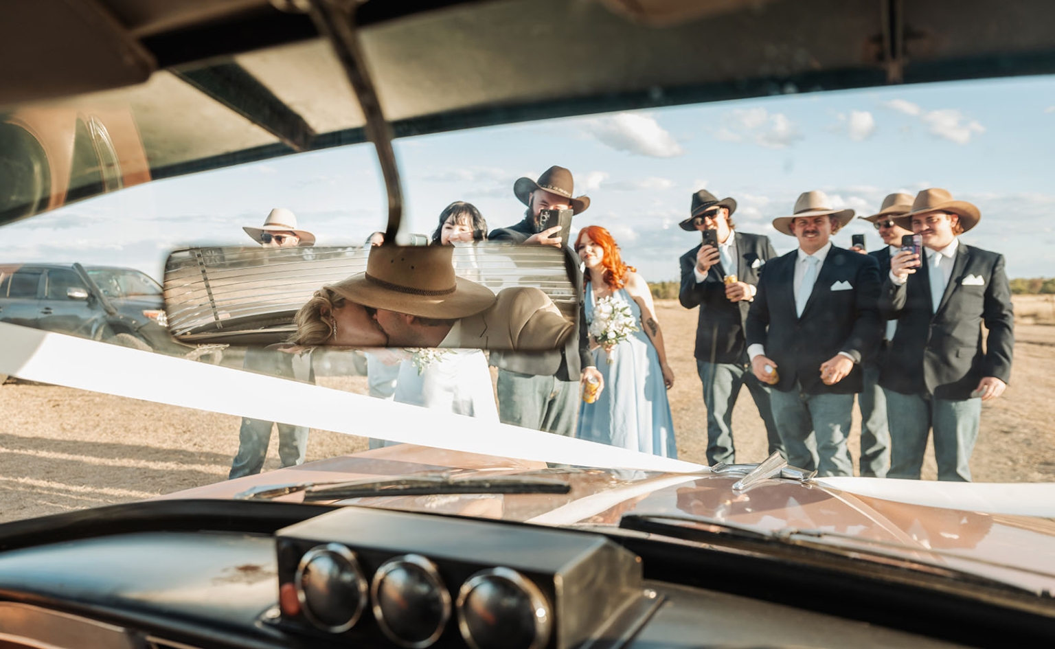 capella wedding photography – caitlin & adam’s farm wedding 159 Newlyweds kissing beside a vintage car as their cowboy-hatted wedding party watches on a farm