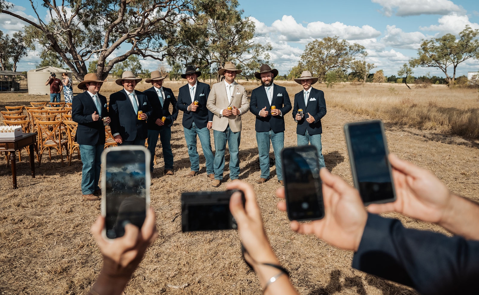 capella wedding photography – caitlin & adam’s farm wedding 136 Wedding party in cowboy hats, jackets and jeans holding drinks as guests photograph them on a dry farm paddock in Capella.