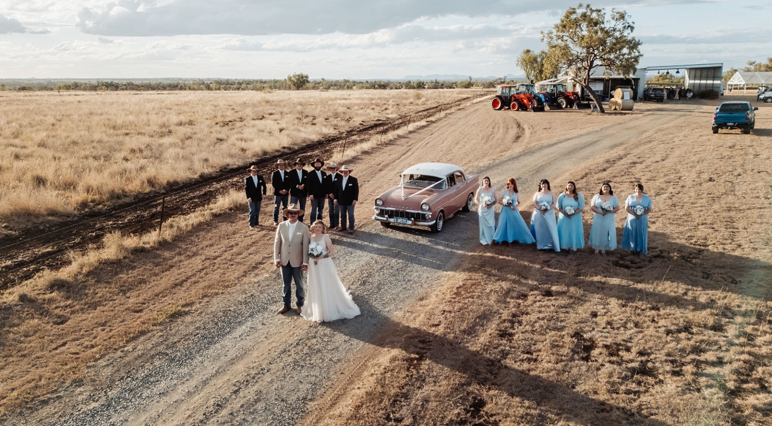capella wedding photography – caitlin & adam’s farm wedding 156 Aerial view of Caitlin and Adam with their wedding party beside a pink vintage car on a dirt road at their Capella farm wedding