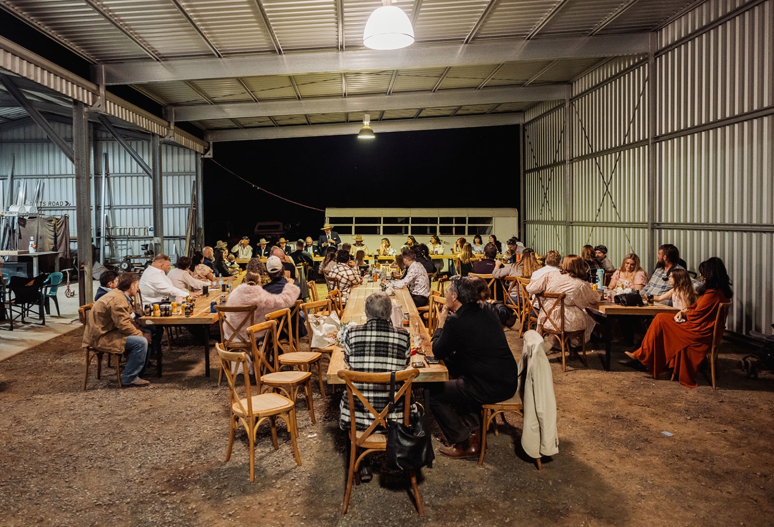 capella wedding photography – caitlin & adam’s farm wedding 181 Wedding guests seated at long tables during an evening reception in a corrugated metal farm shed