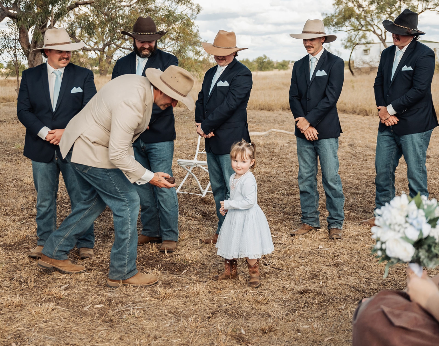 capella wedding photography – caitlin & adam’s farm wedding 147 Man in a cream jacket and cowboy hat bending towards a flower girl as groomsmen in cowboy hats stand behind them at an outdoor farm wedding.
