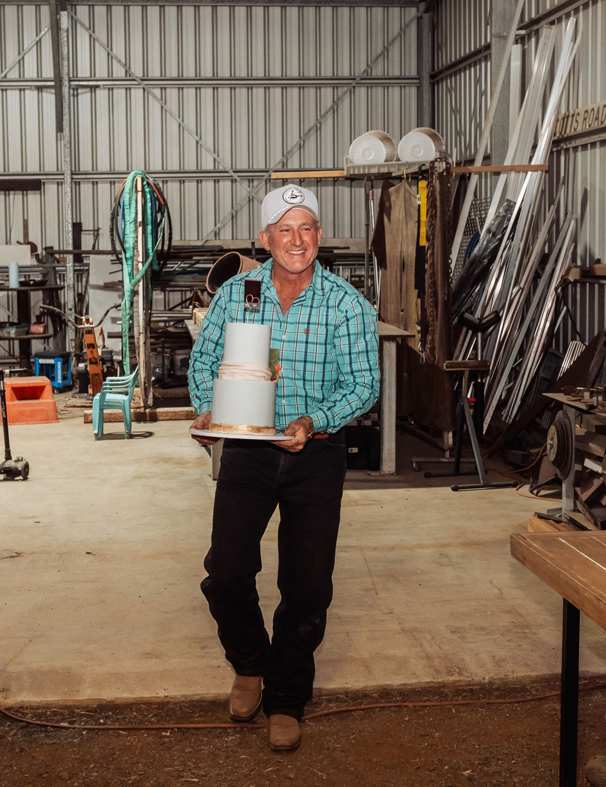 capella wedding photography – caitlin & adam’s farm wedding 185 Man in a blue checked shirt carrying a two-tier wedding cake inside a farm shed.