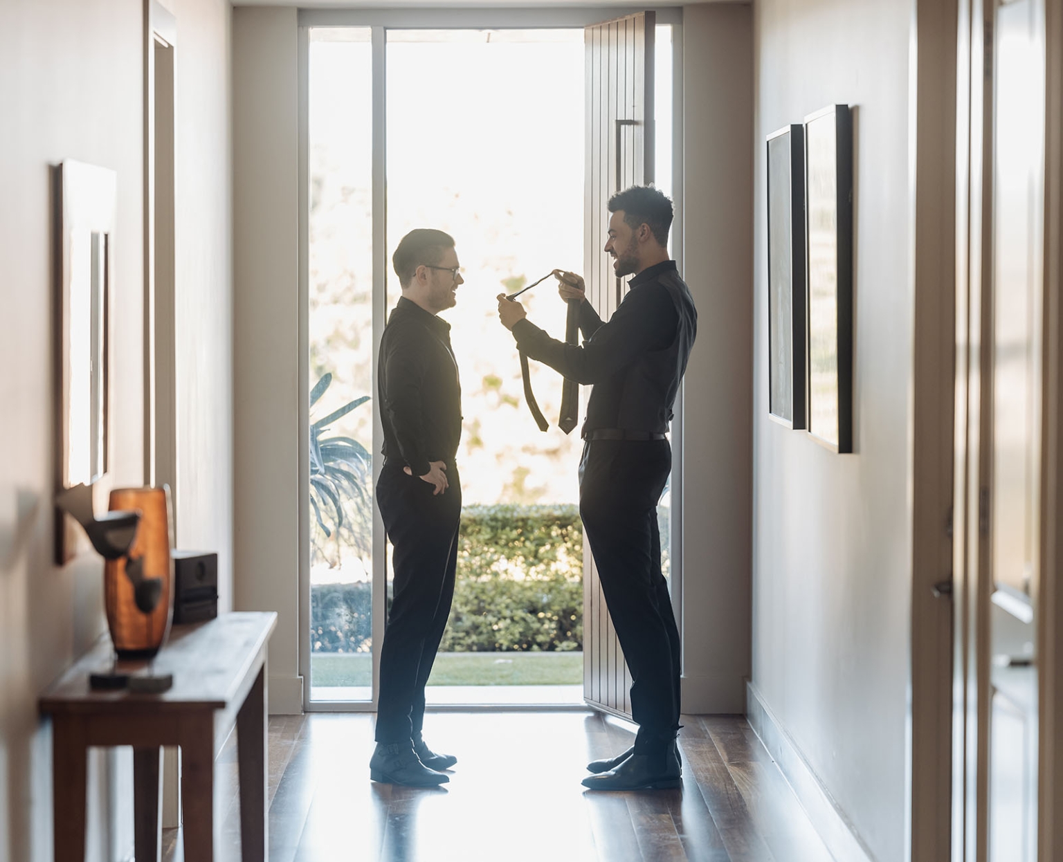 kuitpo hall wedding - courtney & jarrod 193 Two men in formal black suits adjusting a tie in a bright modern hallway.