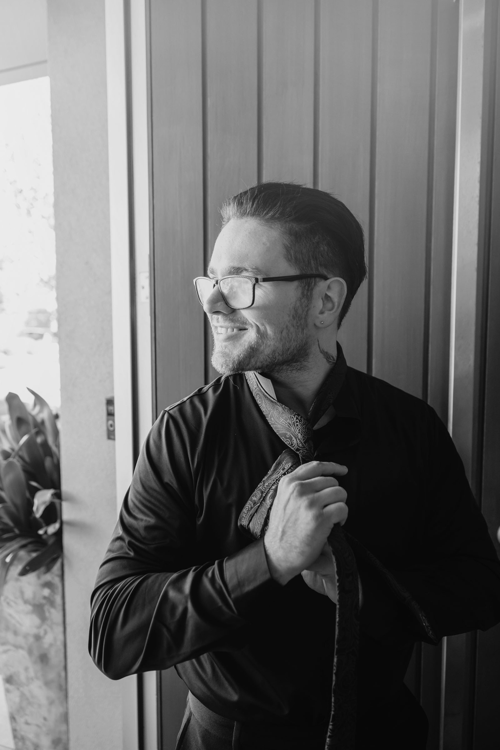 kuitpo hall wedding - courtney & jarrod 195 Professional man adjusting patterned tie by window in dramatic black-and-white portrait.