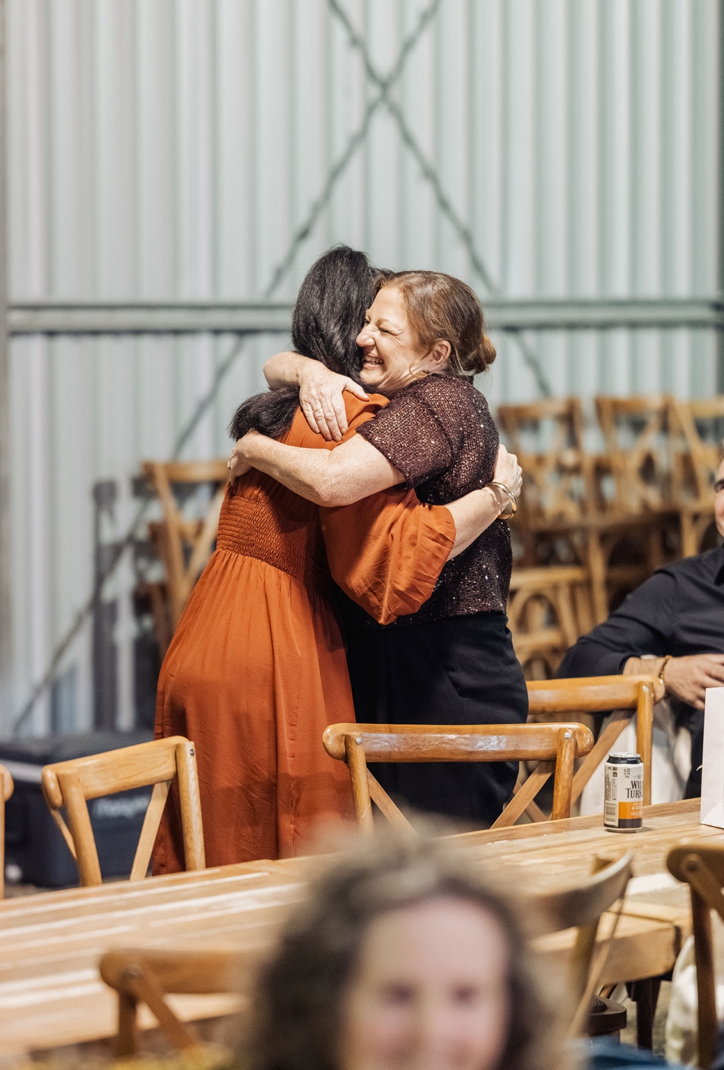 capella wedding photography – caitlin & adam’s farm wedding 183 Two wedding guests embrace joyfully beside wooden tables and chairs at a farm wedding reception.