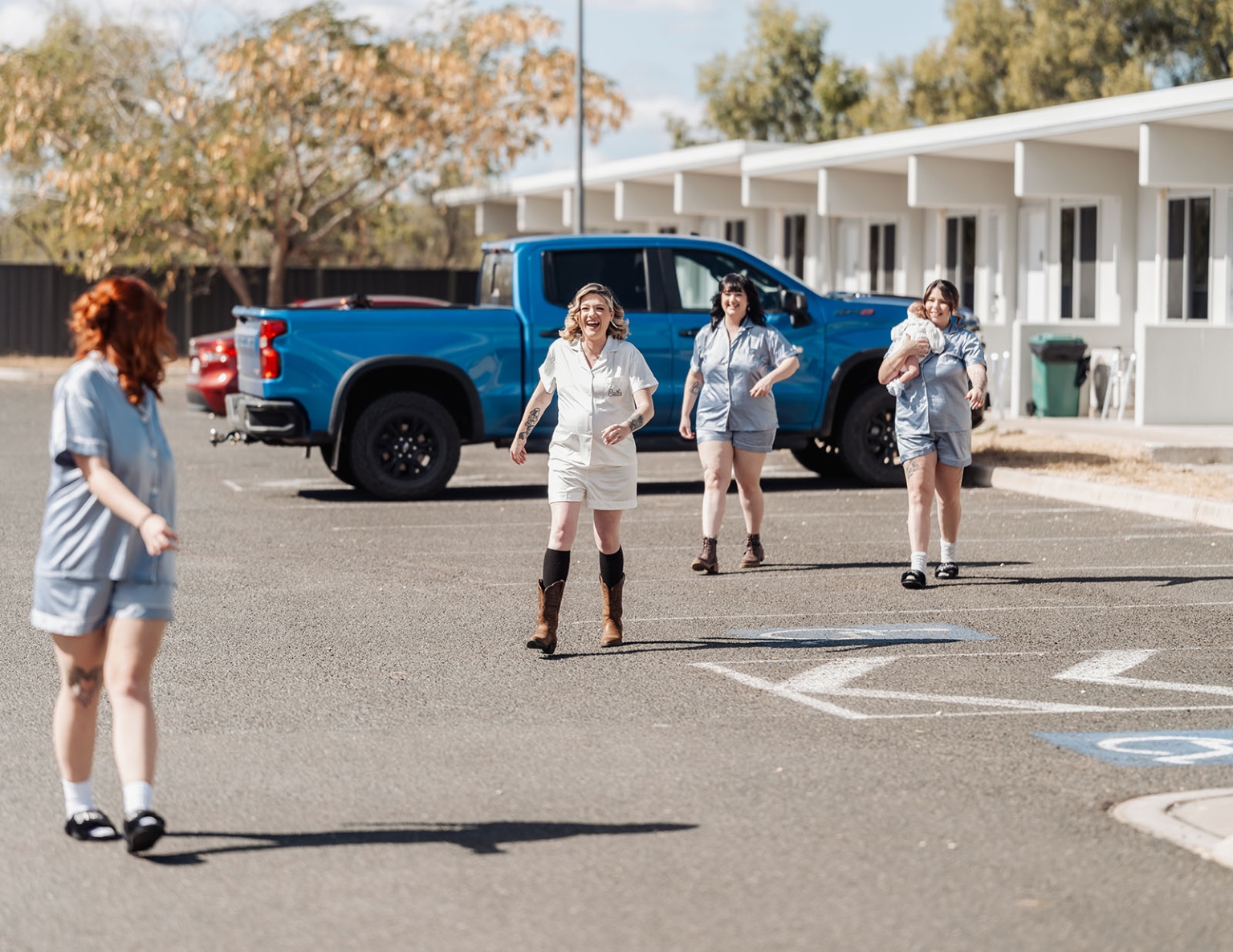 capella wedding photography – caitlin & adam’s farm wedding 123 Women in matching pyjamas walking across a sunny driveway beside a blue pickup truck, with one woman carrying a baby.