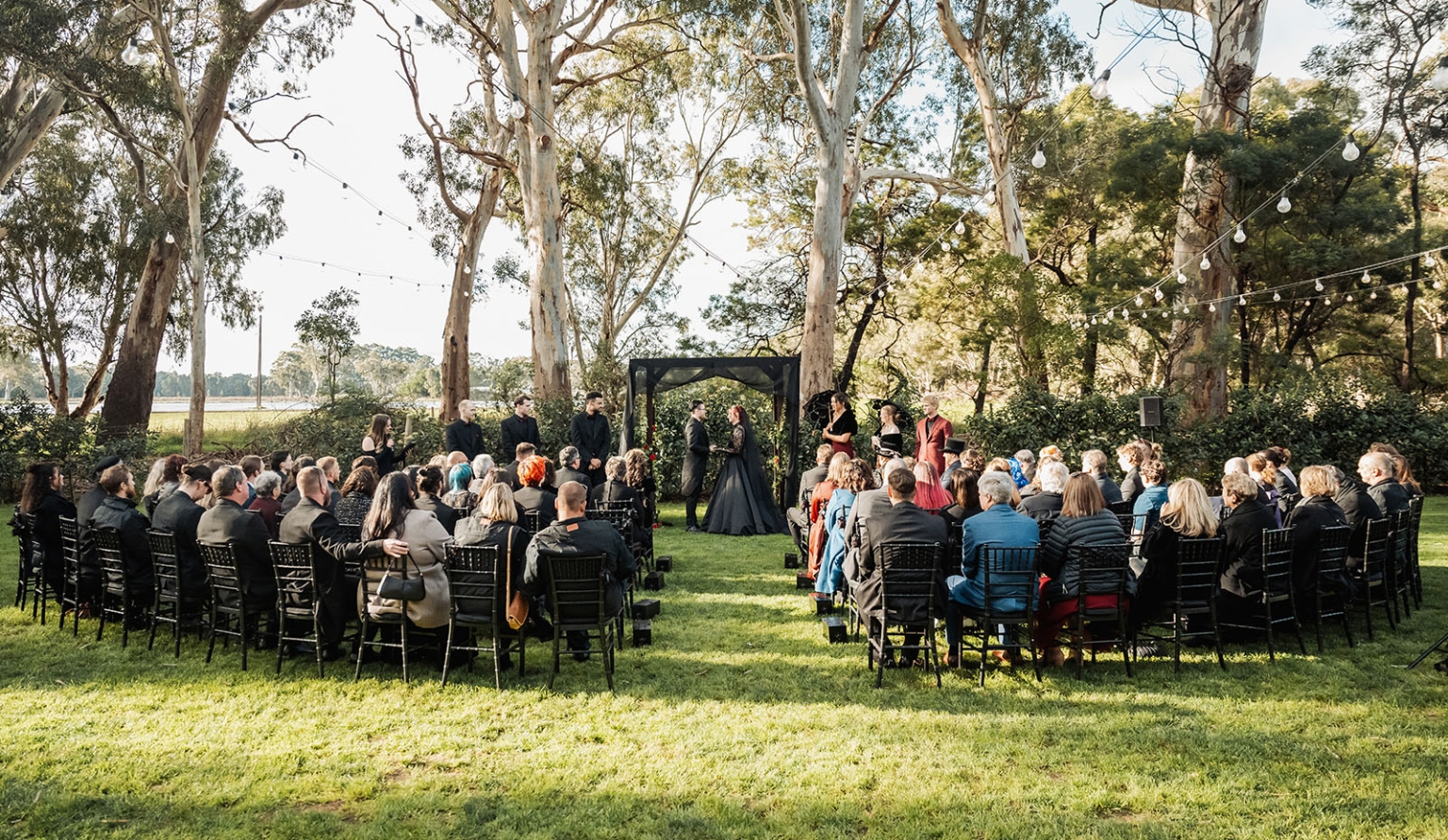 kuitpo hall wedding - courtney & jarrod 225 Gothic woodland wedding ceremony with black decor, outdoor arch, seated guests, and string lights.