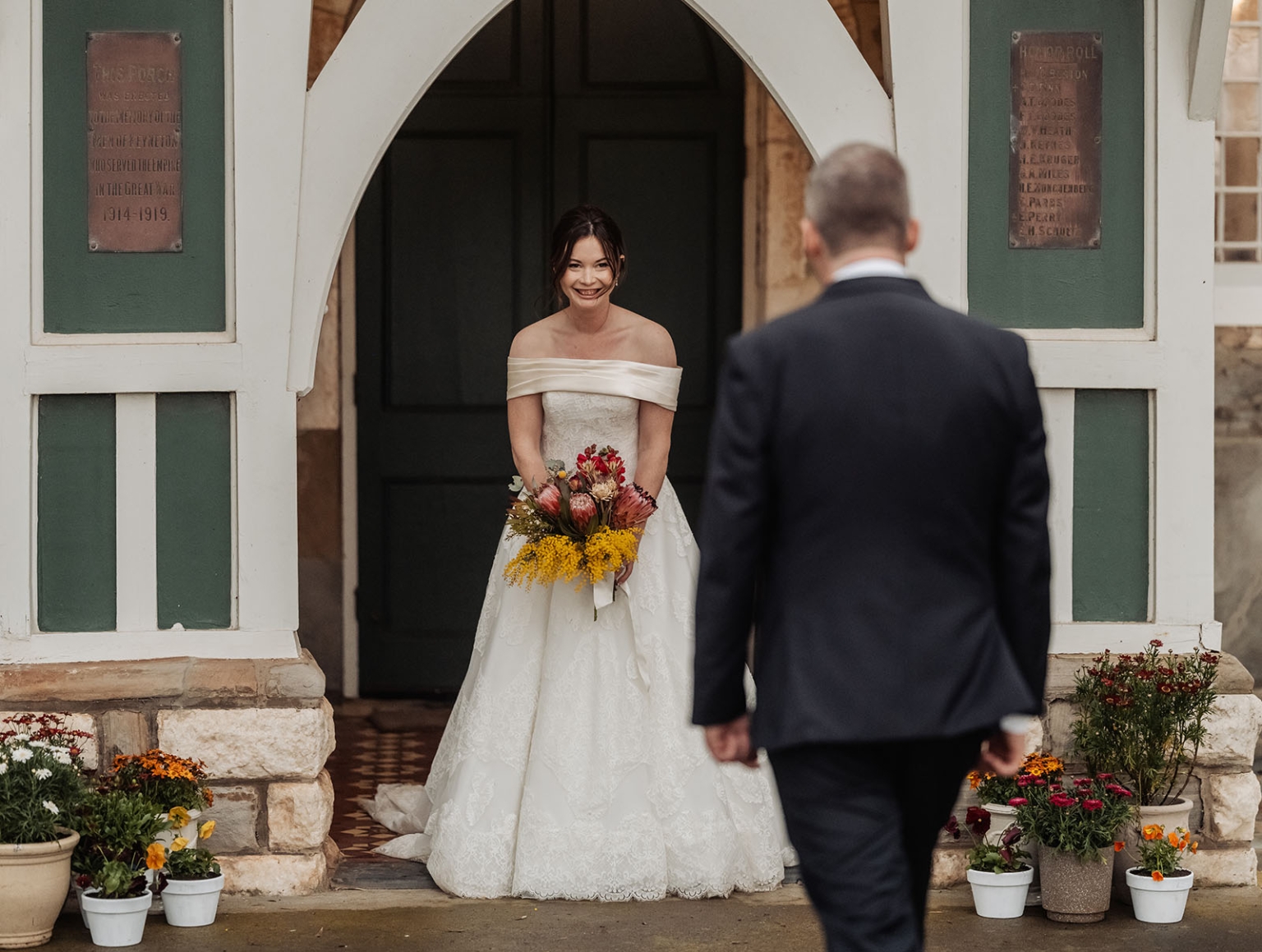 keyneton soldier's memorial hall wedding - lara & simon 29 Bride in an off-the-shoulder wedding dress holding a colourful native flower bouquet as the groom approaches the church entrance.