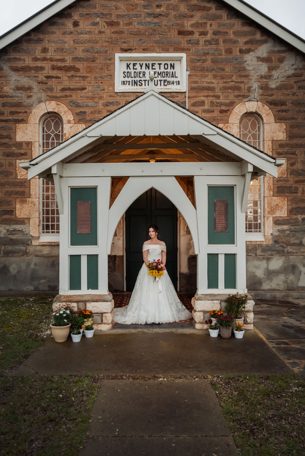 keyneton soldier's memorial hall wedding - lara & simon 30 Bride in an off-the-shoulder white wedding dress holding a colourful bouquet beneath the entrance of Keyneton Soldier's Memorial Hall