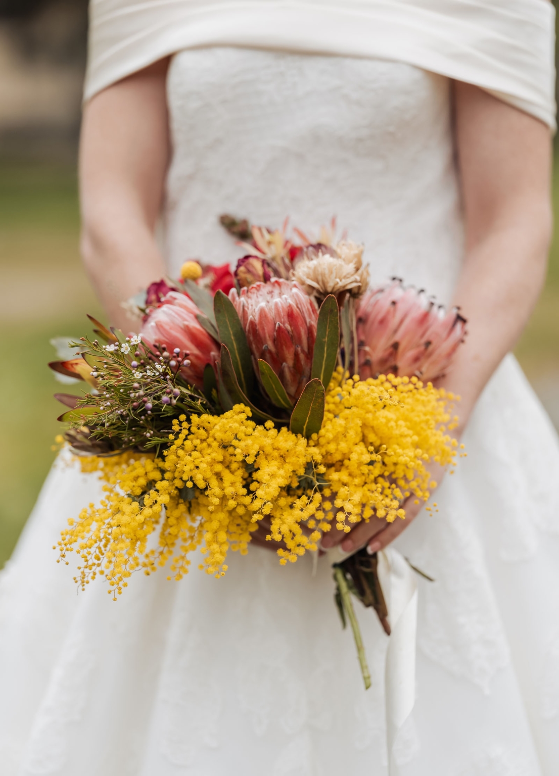 keyneton soldier's memorial hall wedding - lara & simon 21 Bride in an off-the-shoulder white wedding dress holding a bouquet of pink proteas and yellow wattle.
