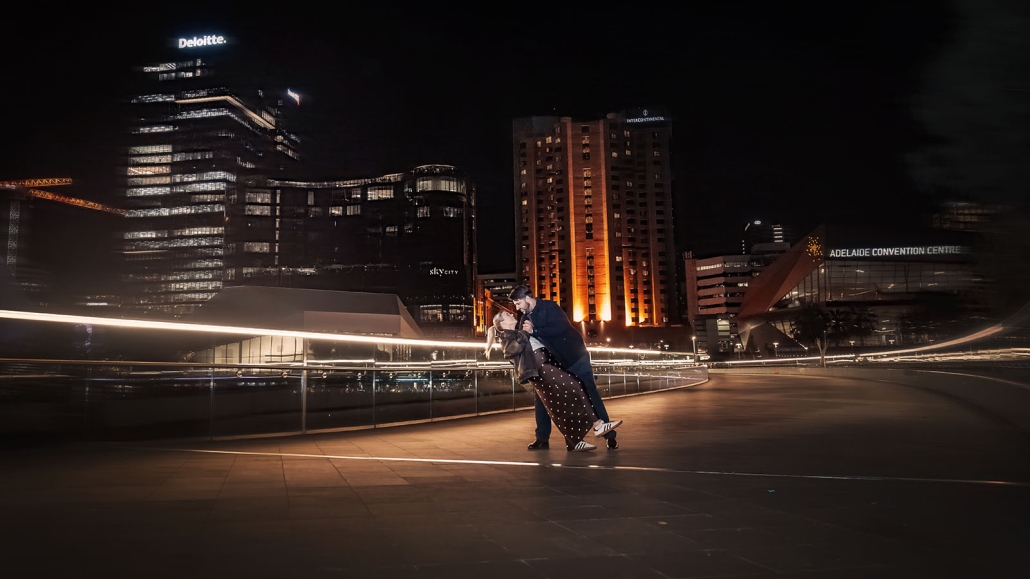 elder park rotunda proposal - lauren & enrique 41 Couple dancing together on the Adelaide riverbank at night, with illuminated city buildings behind them