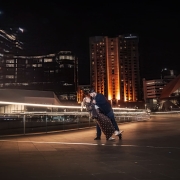 Couple dancing together on the Adelaide riverbank at night, with illuminated city buildings behind them