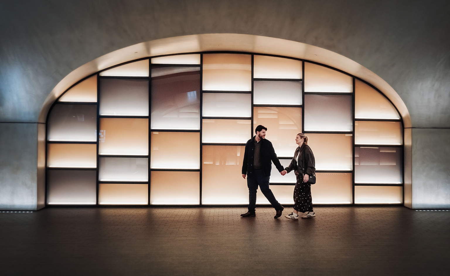elder park rotunda proposal - lauren & enrique 18 Couple holding hands as they walk past a large illuminated arched window at night