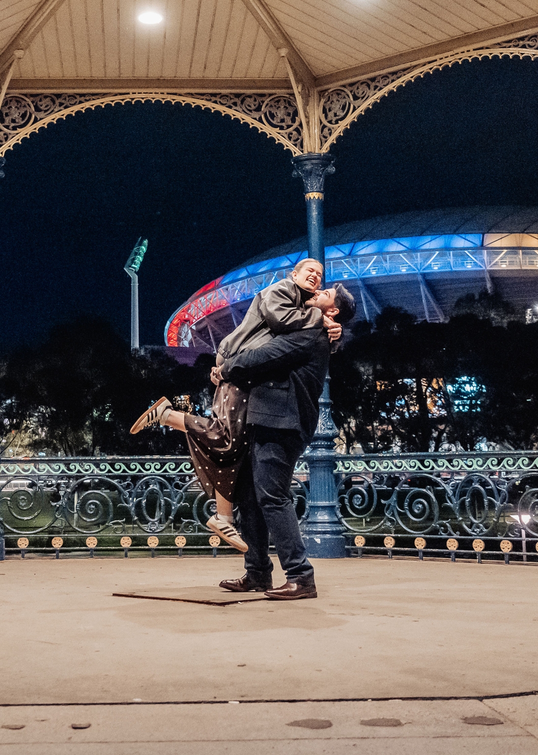 elder park rotunda proposal - lauren & enrique 10 Lauren and Enrique embracing as he lifts her beneath the Elder Park Rotunda at night, with Adelaide Oval lit behind them