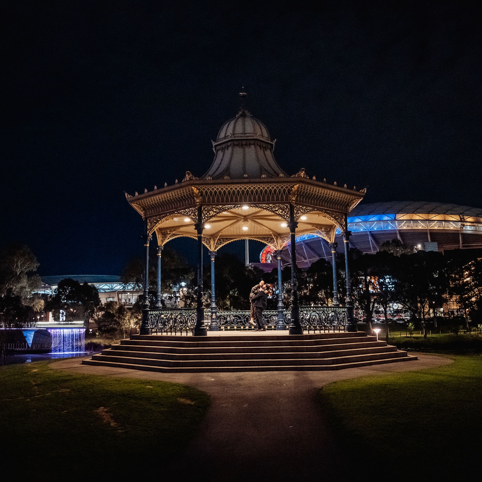 elder park rotunda proposal - lauren & enrique 9 Lauren and Enrique embracing beneath the illuminated Elder Park Rotunda at night in Adelaide