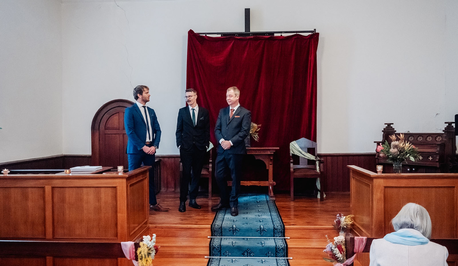 keyneton soldier's memorial hall wedding - lara & simon 5 Three men in suits stand at the front of Keyneton Church during a wedding ceremony, beneath a burgundy curtain covering the cross.