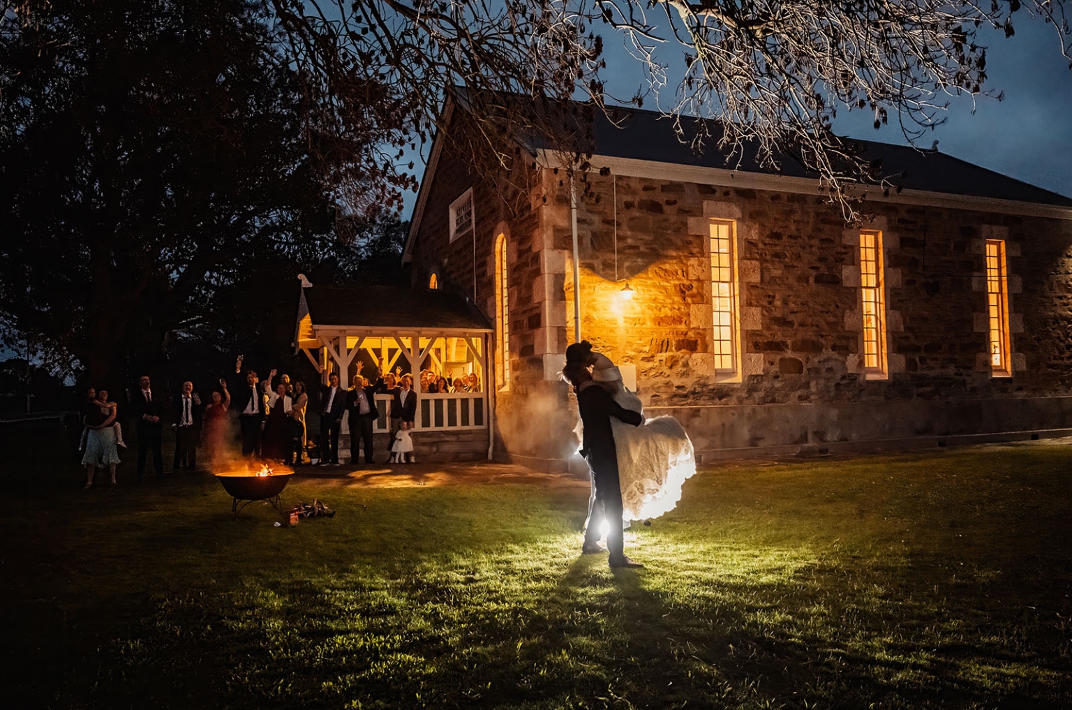 keyneton soldier's memorial hall wedding - lara & simon 47 Newlyweds embrace on the lawn outside Keyneton Church at night as wedding guests watch beside a fire pit.