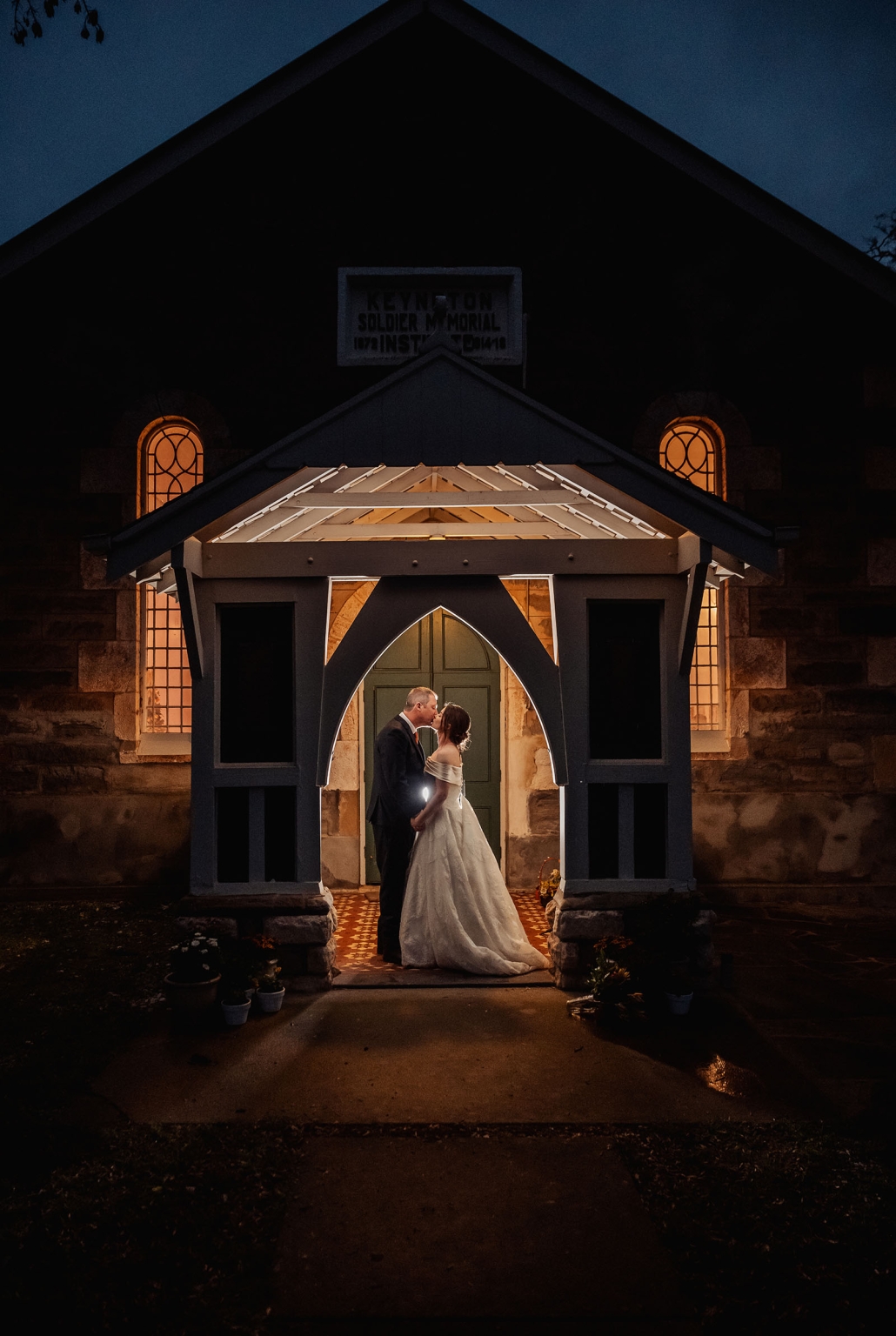 keyneton soldier's memorial hall wedding - lara & simon 46 Bride and groom kissing beneath the illuminated entrance of Keyneton Soldier's Memorial Hall at night
