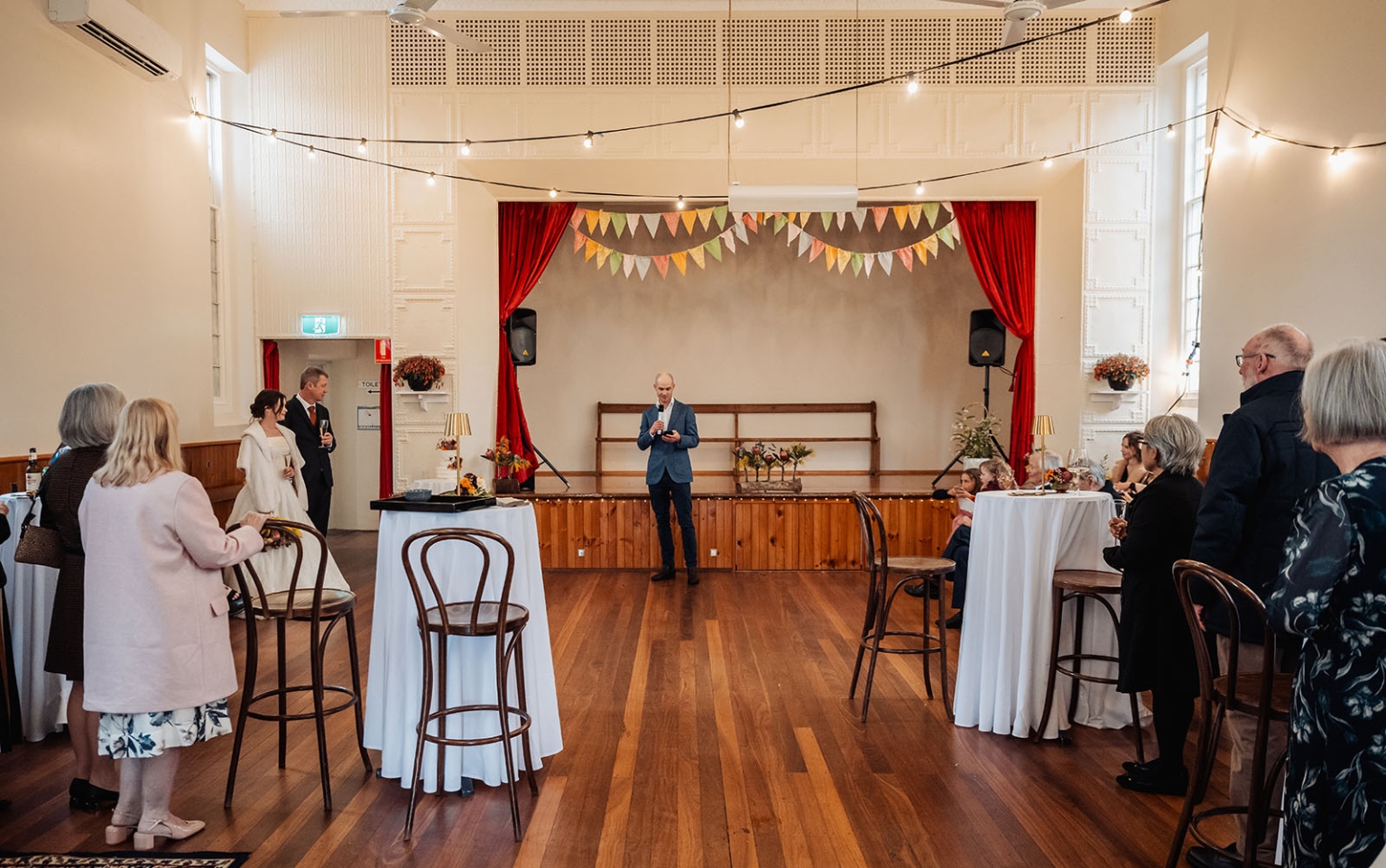 keyneton soldier's memorial hall wedding - lara & simon 42 Wedding guests gathered in Keyneton Soldier's Memorial Hall as a man gives a speech near the stage, with the couple standing beside him.
